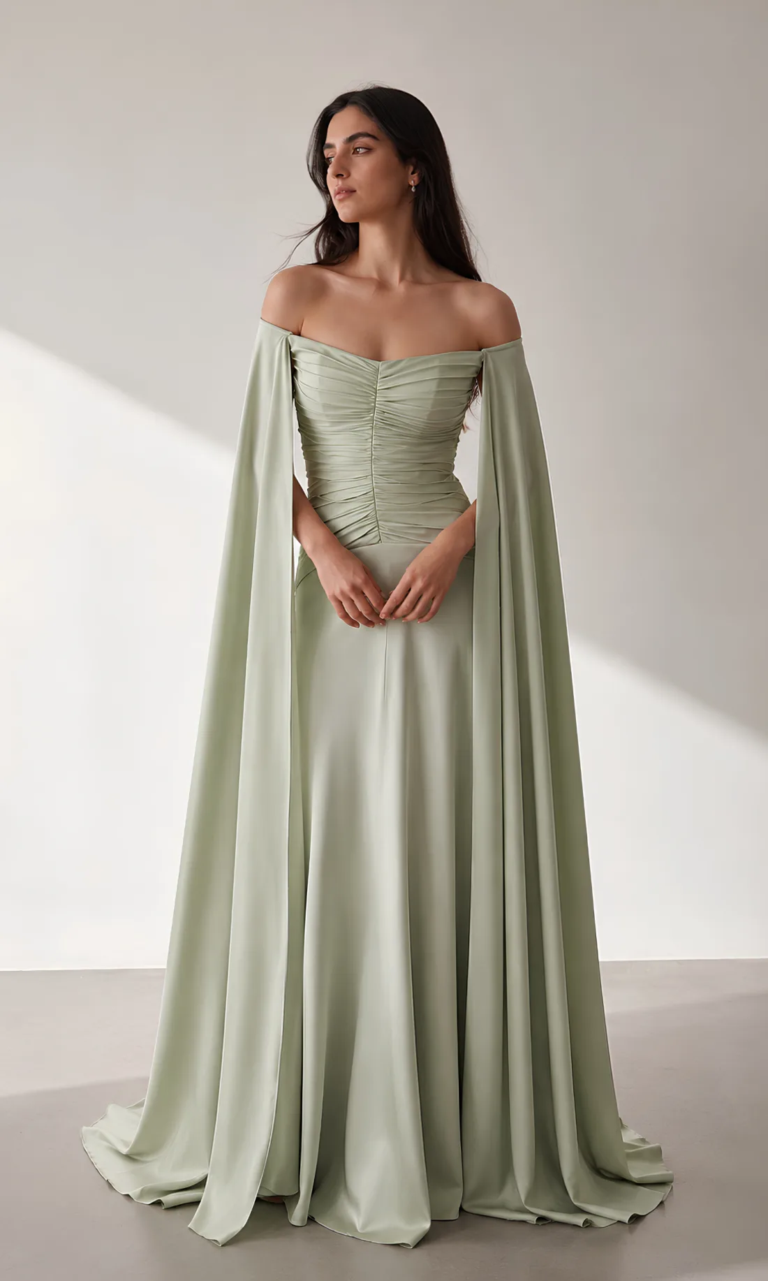A model wearing the Joie Couture sage green off-shoulder gown with long draped cape sleeves, photographed against a pale studio ground